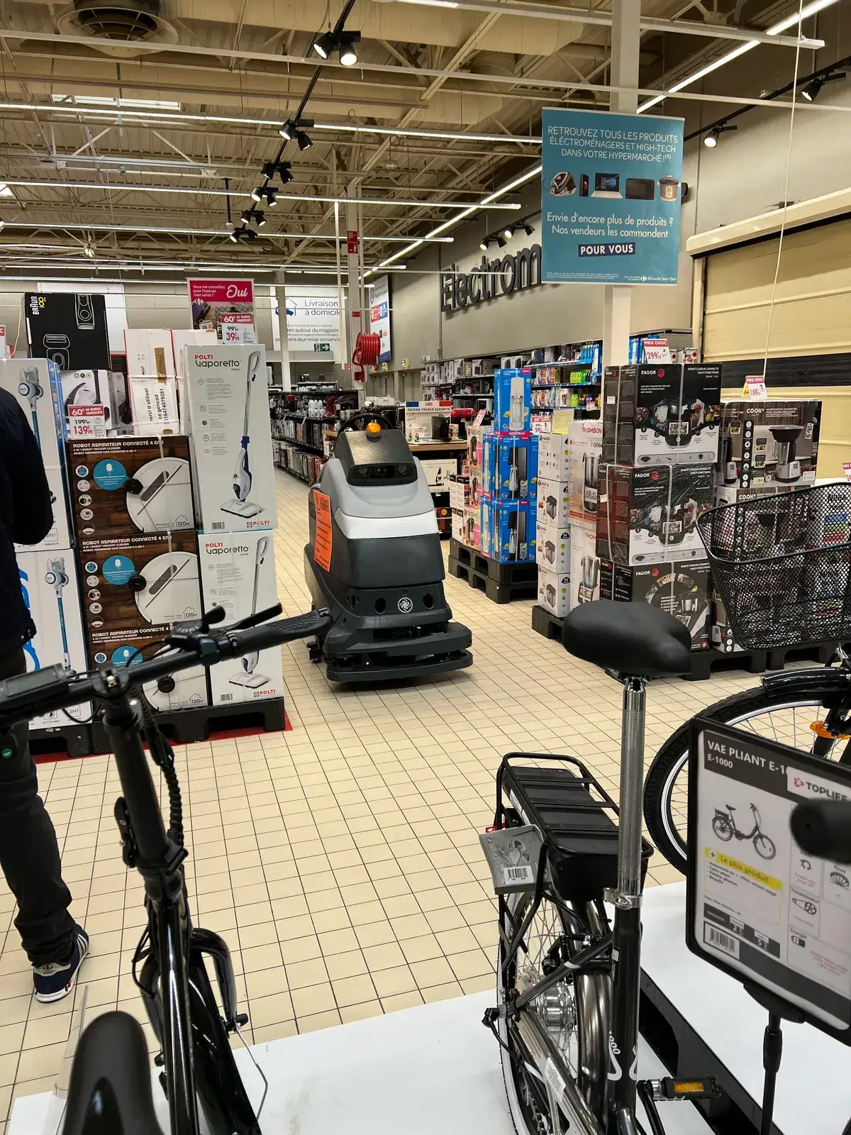 A large autonomous floor-scrubbing robot parked in the appliance aisle of a Carrefour hypermarket