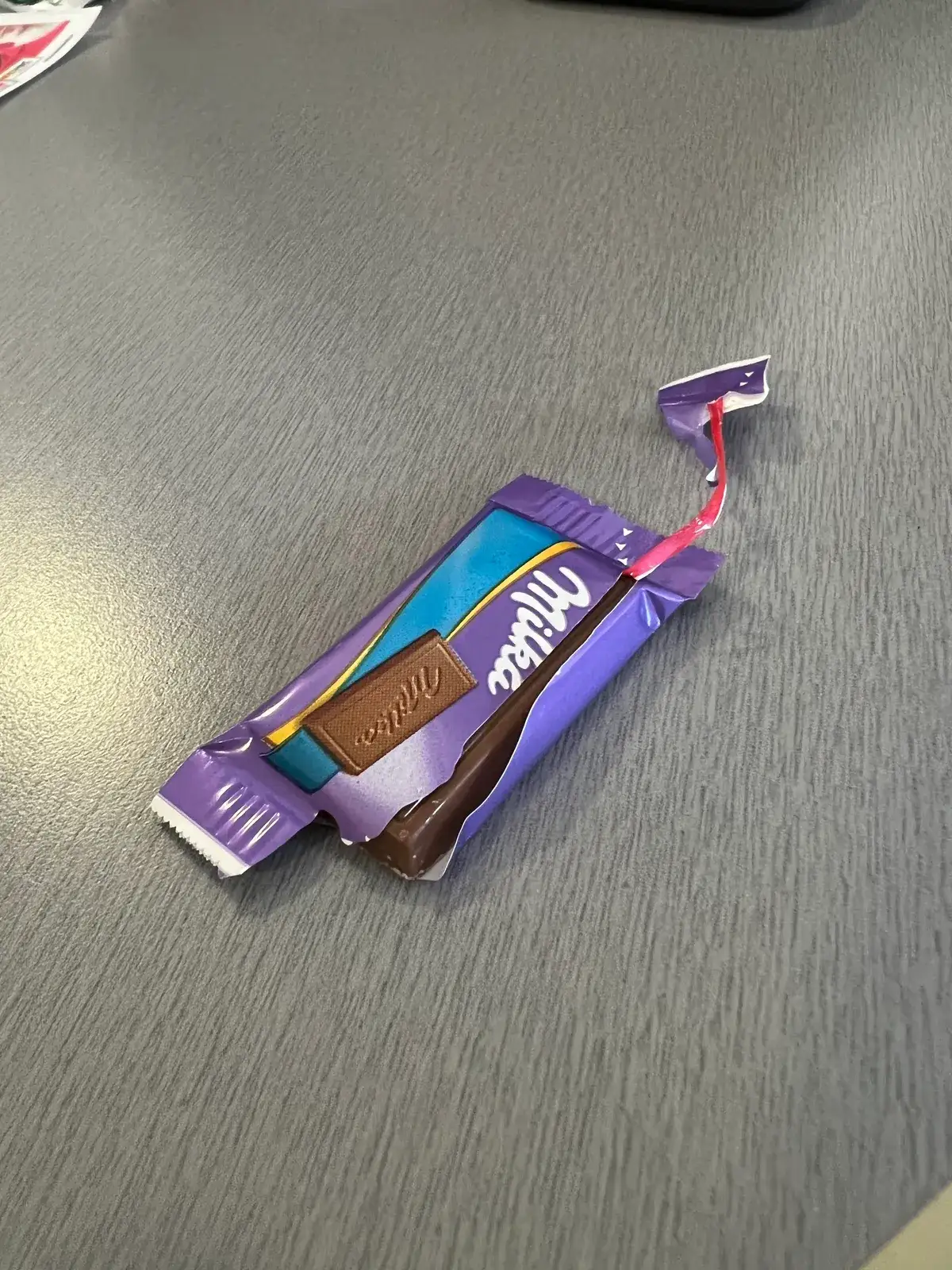 A small Milka chocolate bar on a desk, its wrapper opened by a red tear strip pulled halfway along the package