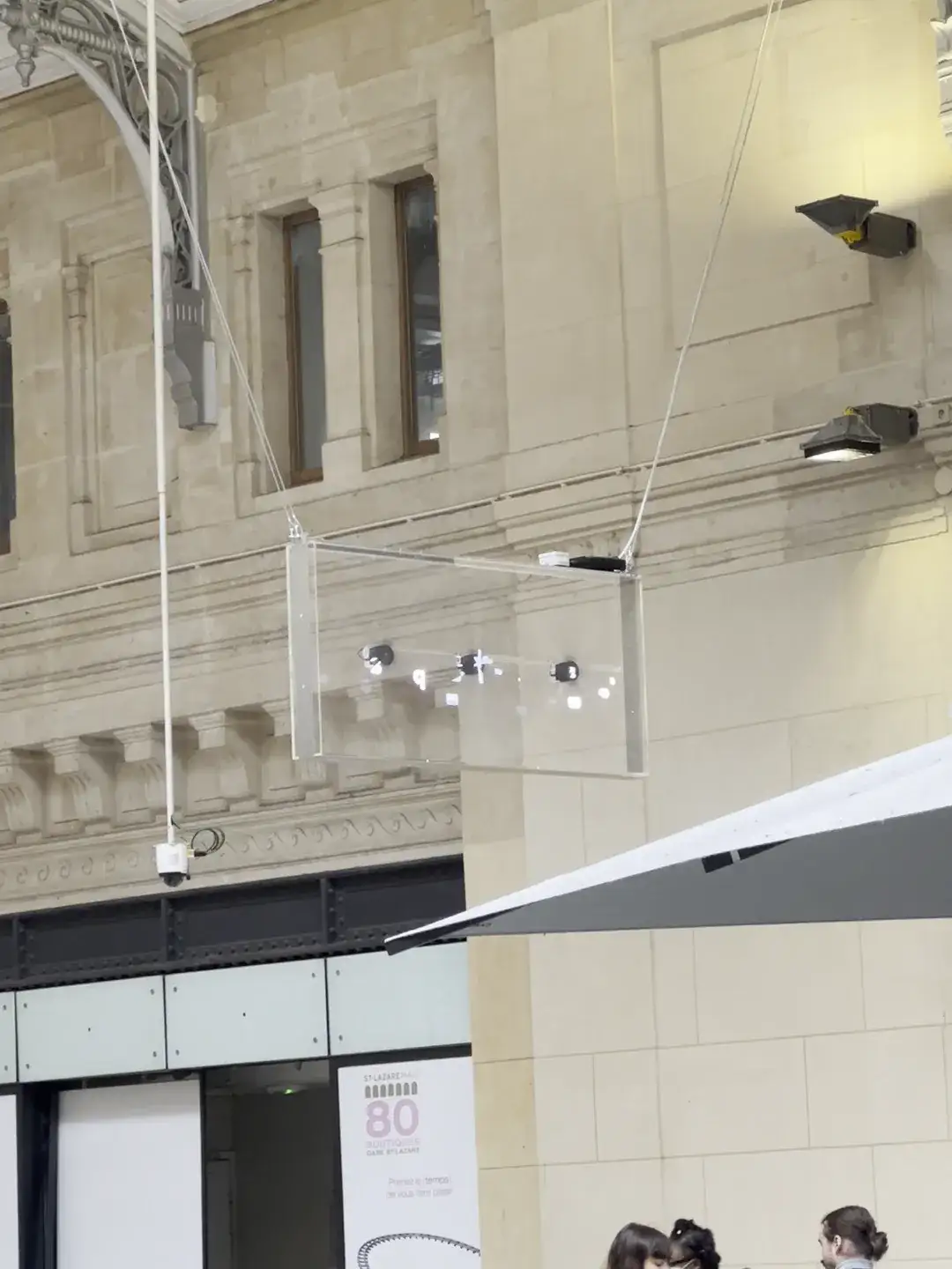 A holographic LED fan display suspended on cables from the stone wall of the Gare Saint-Lazare concourse, spinning above café tables of seated travellers