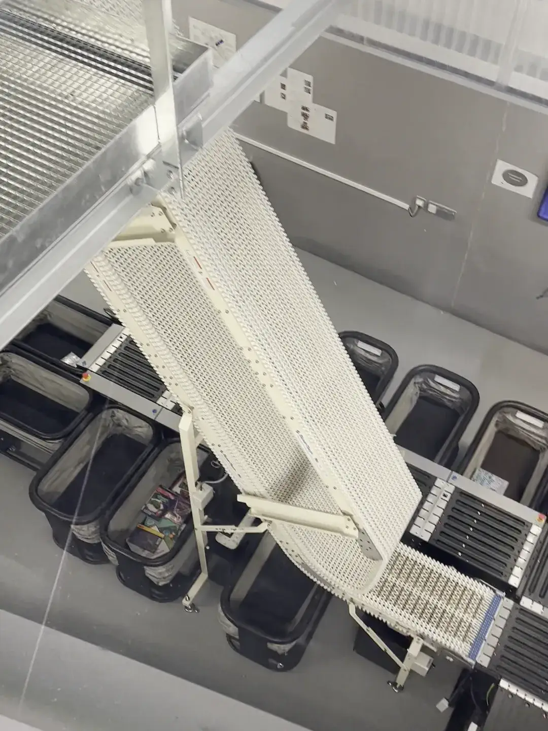 Looking down through glass at an automated book-return system in the Library of Caen, where a white modular conveyor belt curves down to drop returned books into black plastic bins