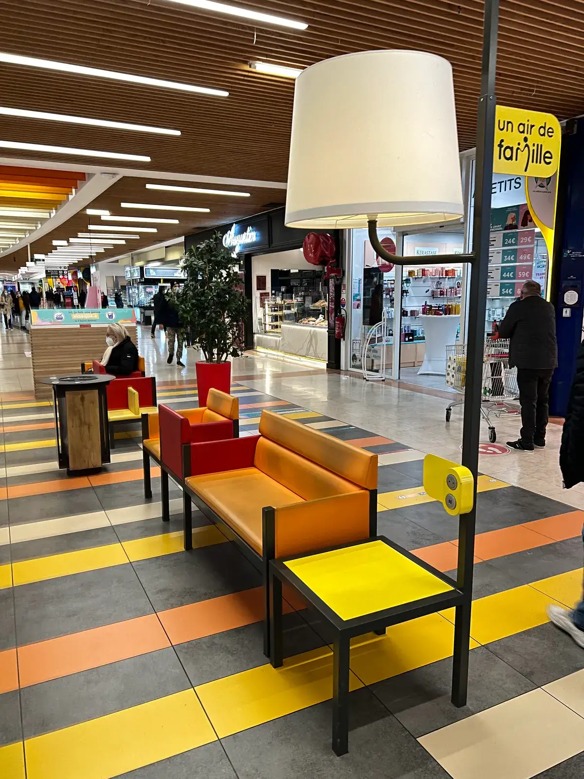 A mall corridor lounge with orange and red benches, an oversized lamp with built-in power outlets, and a floor of yellow, orange and grey stripes