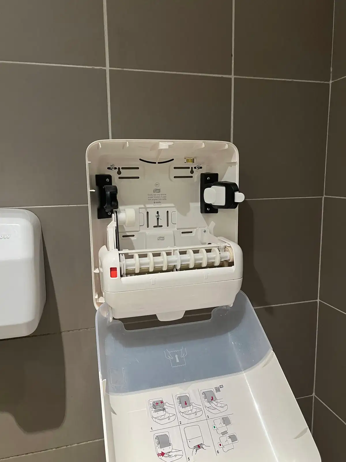 A wall-mounted Tork paper towel dispenser hanging open, showing the bubble level in its backplate, the refill mechanism, and illustrated reloading instructions on the cover