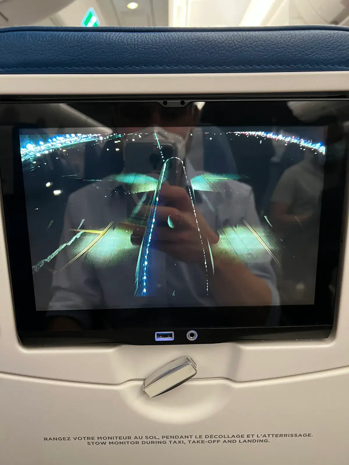 Seat-back screen on an airplane showing a live bird's-eye camera view of the wing and runway lights at night