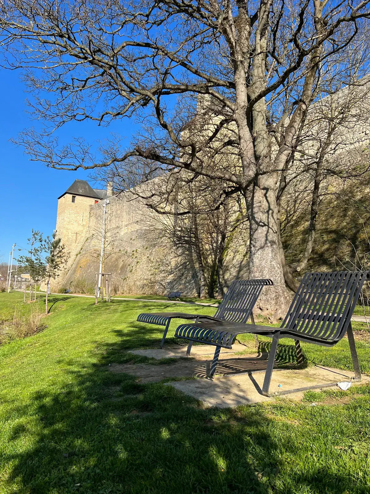 Two slatted metal loungers on a concrete pad in the grass below the stone ramparts of the Château de Caen