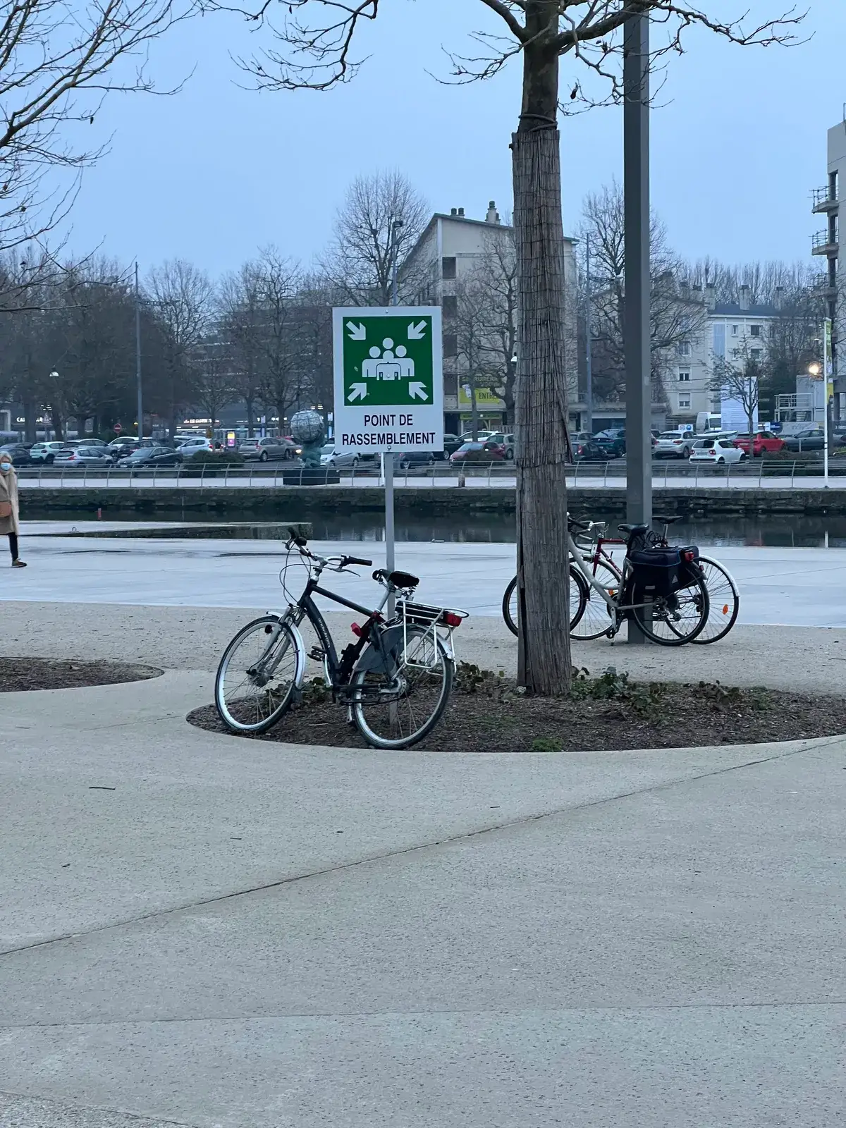 A green POINT DE RASSEMBLEMENT sign on a post beside two parked bicycles, on a waterfront plaza