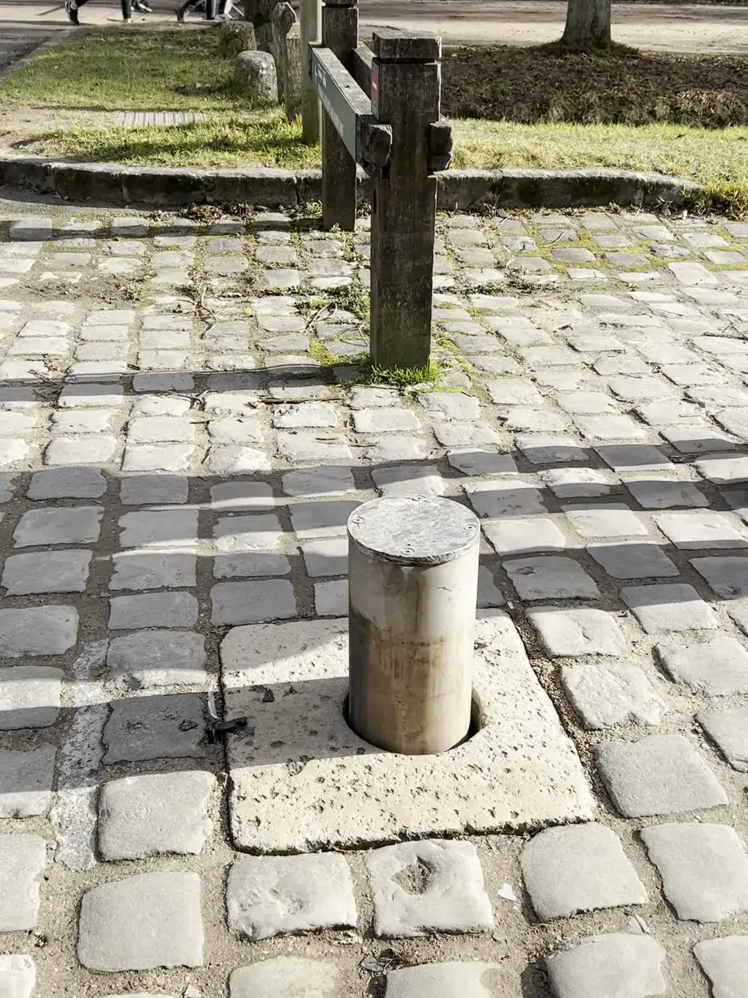A metal retractable bollard set into a cobblestone forecourt at the Château de Versailles, with wooden posts, bare winter trees and passing walkers behind it