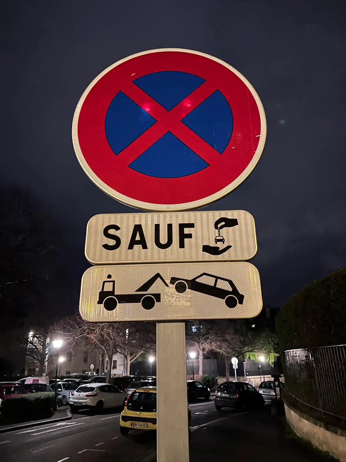 A no-stopping sign at night with two plates beneath it: SAUF next to a hand pictogram, and a tow truck hauling a car away