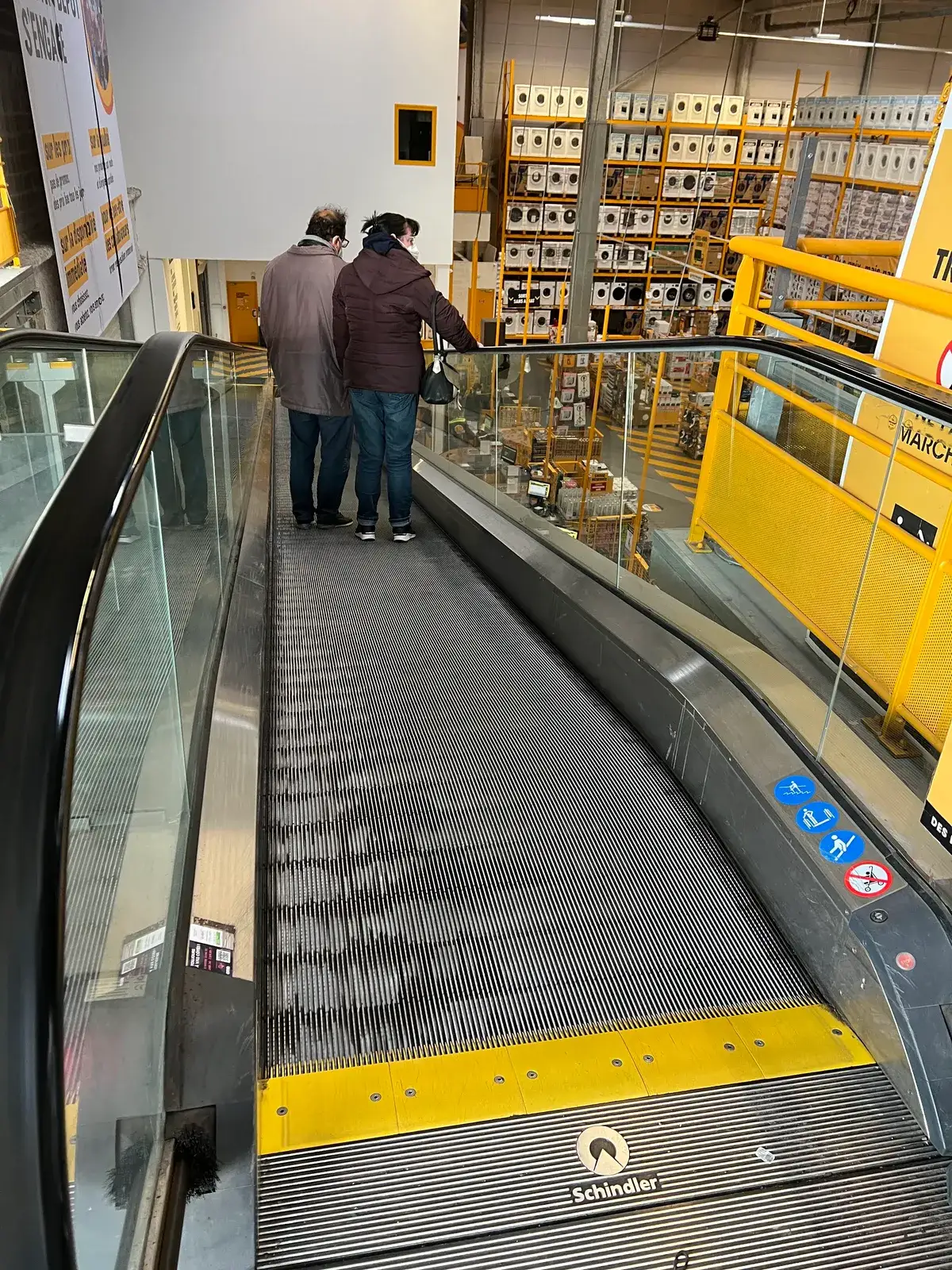 A stepless inclined moving walkway carrying two shoppers down past shelves stacked with washing machines