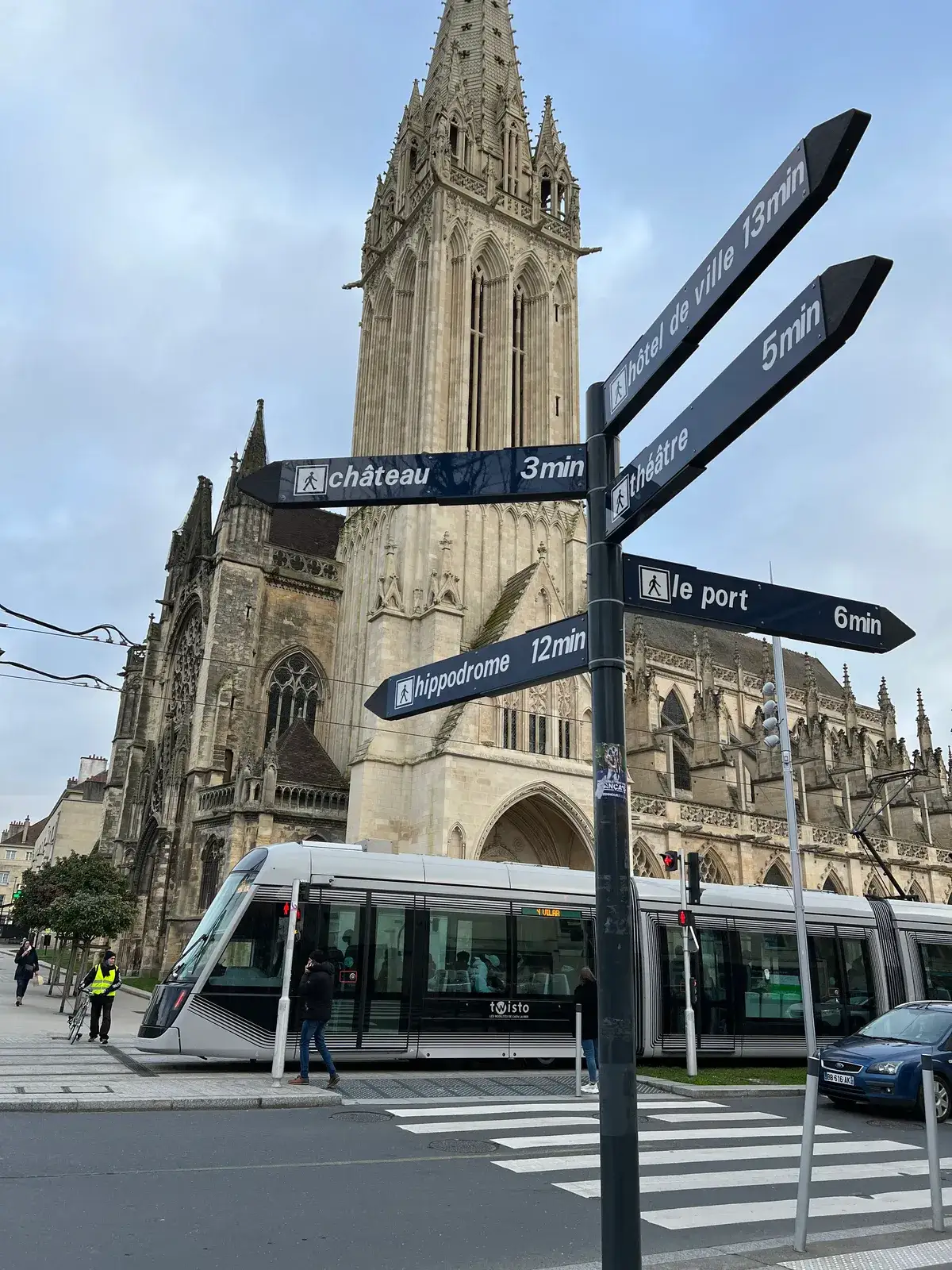 A signpost of dark blue pedestrian fingerposts — château 3min, le port 6min, hôtel de ville 13min, hippodrome 12min — in front of a Gothic church and a passing tram
